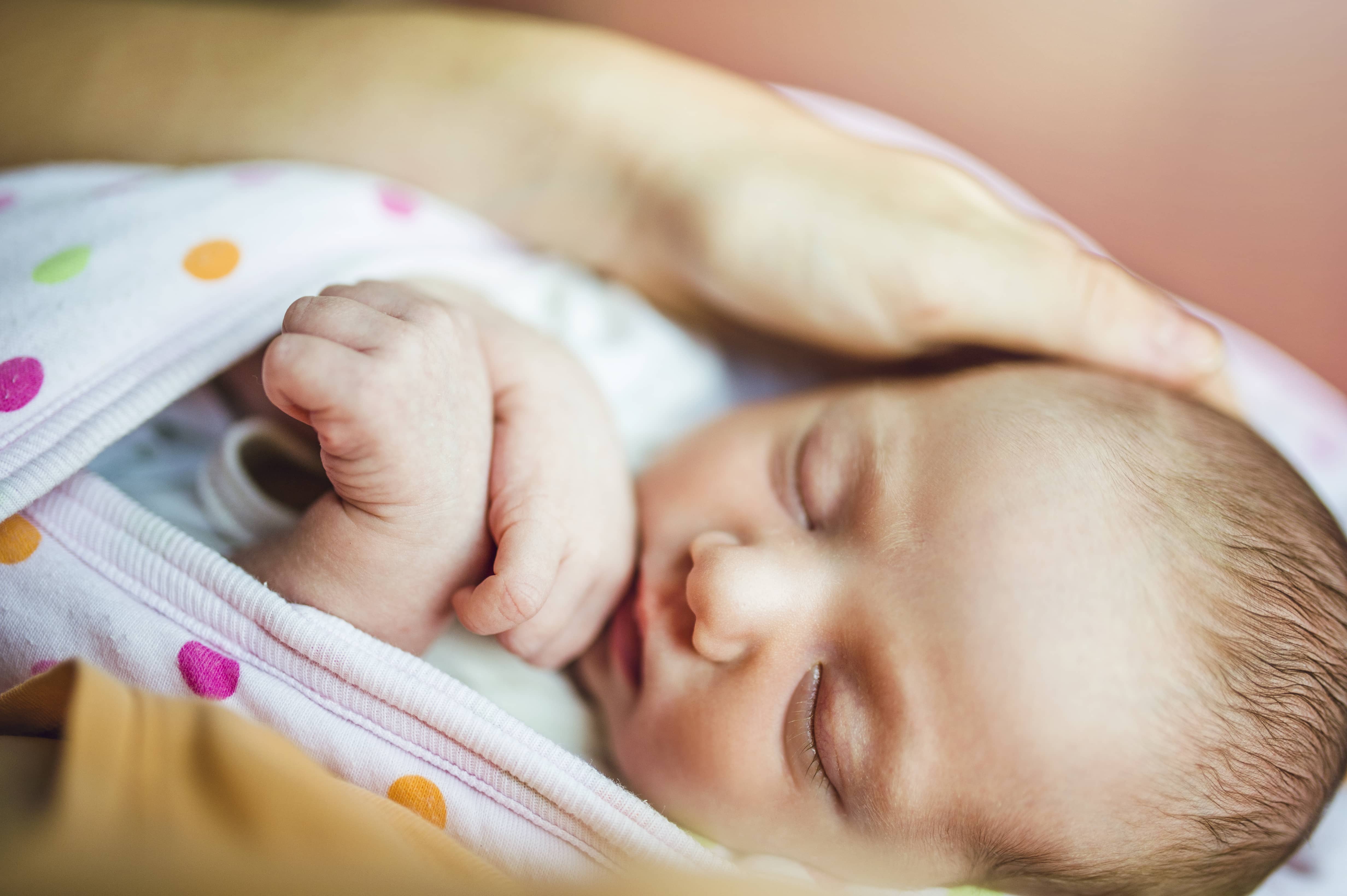 Sleeping tucked in baby in a blanket with an adult hand resting on the baby's head